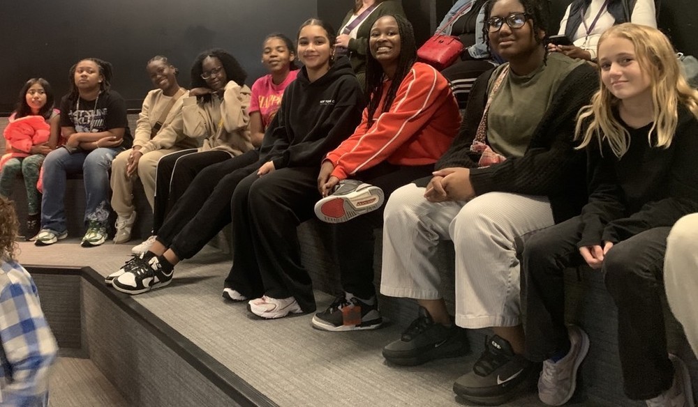 A group of students seated on a bench, facing a large crowd gathered in front of them.