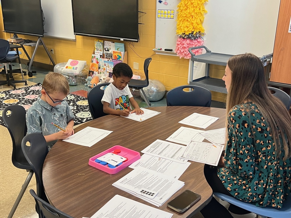 To students working on worksheets at a table with the teacher sitting before them.