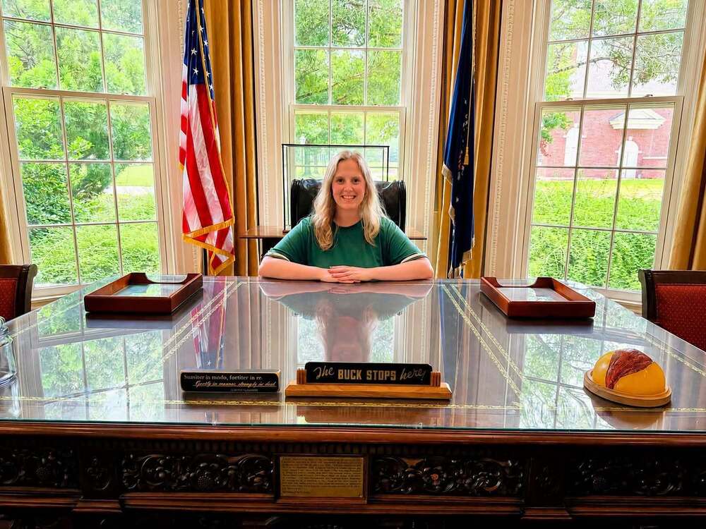 Mrs. Frank sitting behind a desk with the American Flag to her left and another flag to her right. There are three windows behind her.
