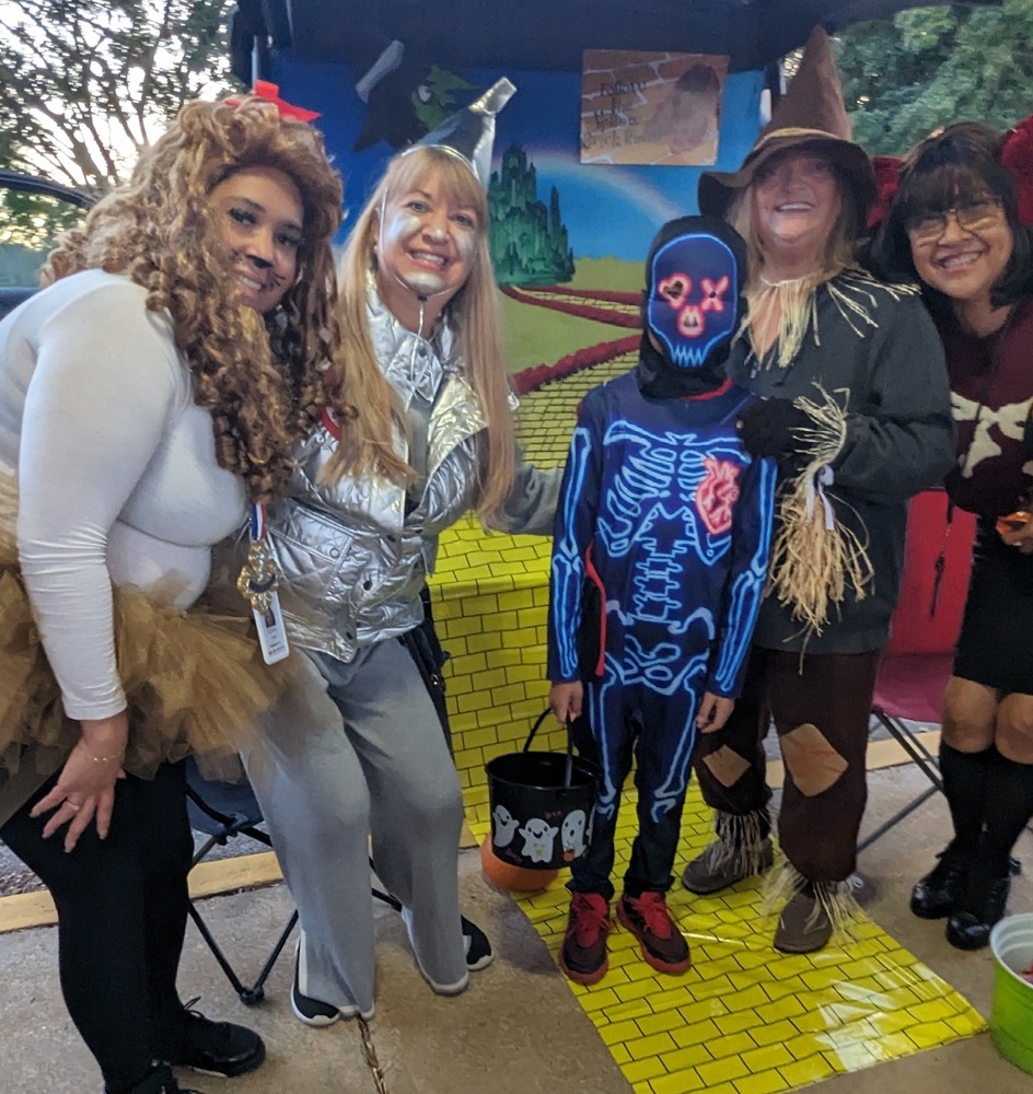 Highlands teachers dressed in costumes pose with a student wearing a fun costume during the PTA Trunk or Treat event.