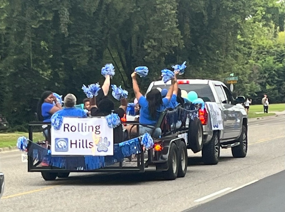 A parade float decorated in blue with pom-poms, balloons, and a banner reading “Rolling Hills” is pulled by a pickup truck. A few Rolling Hills staff members wearing blue shirts ride on the float, cheering and waving pom-poms along the parade route.
