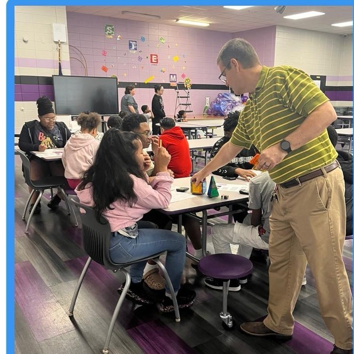 A group of students are sitting at tables in the cafeteria with different shapes on the table. Their teacher stands at the table with them