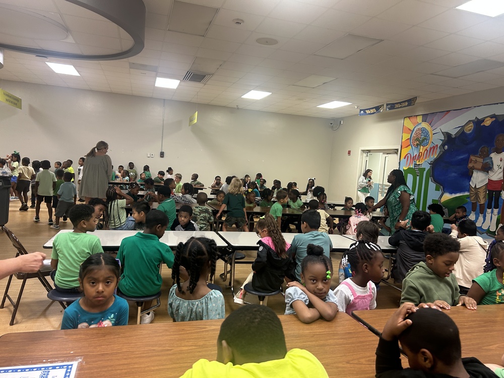 Students and staff at RHES gather in the cafeteria dressed in green outfits for Color Days. Children are seated at tables, while some are standing and talking. Teachers and staff walk among the groups, and a colorful mural with the words “Dream Big” is visible on the wall in the background.