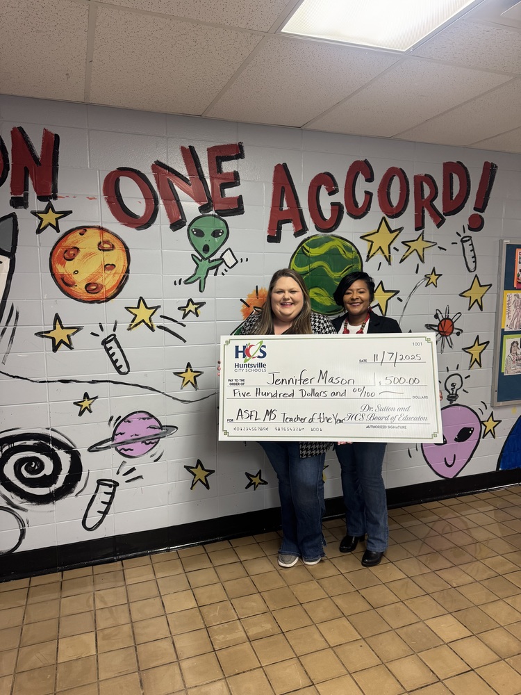 Collaborative teacher and Principal Shoulders proudly display a large check of $500 in front of a textured wall.