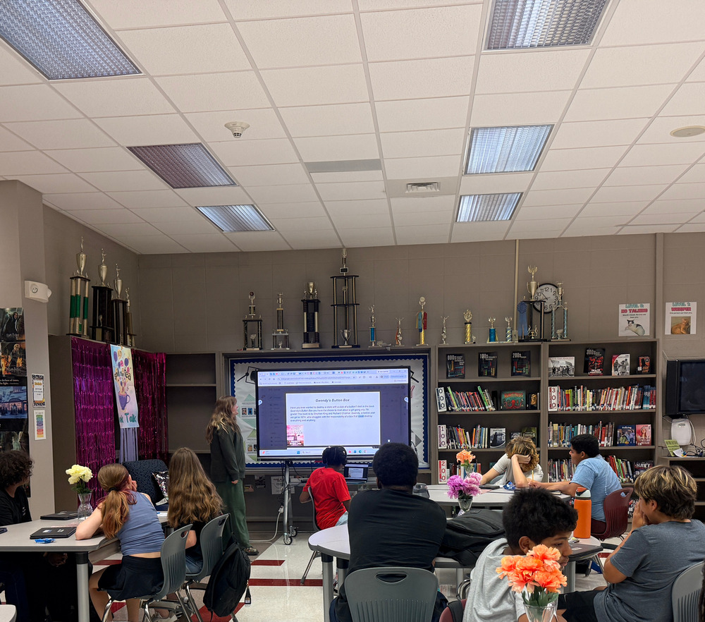 8th grade students sitting and listening in library as student presents book talk