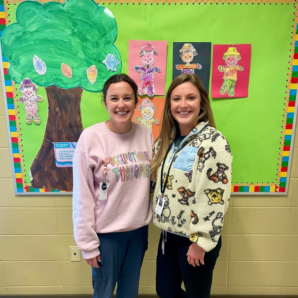 Two Goldsmith Occupational Therapists stand in front of a fall theme bulletin board with a tree and scarecrows