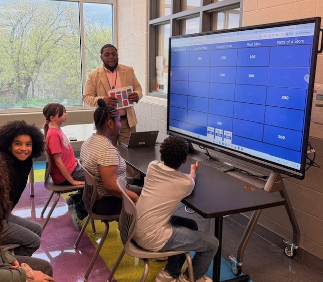 Assistant Principal Irving McGuire stands beside a large interactive screen displaying a Jeopardy-style review game as part of a “Moving to Mastery” station focused on system principles. Several students, identified as McKenzie Yeager, Zechariah Conner, Joi Desouza are seated around a table, actively engaged in the activity. One student points at the board while others watch and participate, demonstrating collaboration, critical thinking, and excitement for learning in a small-group instructional setting.