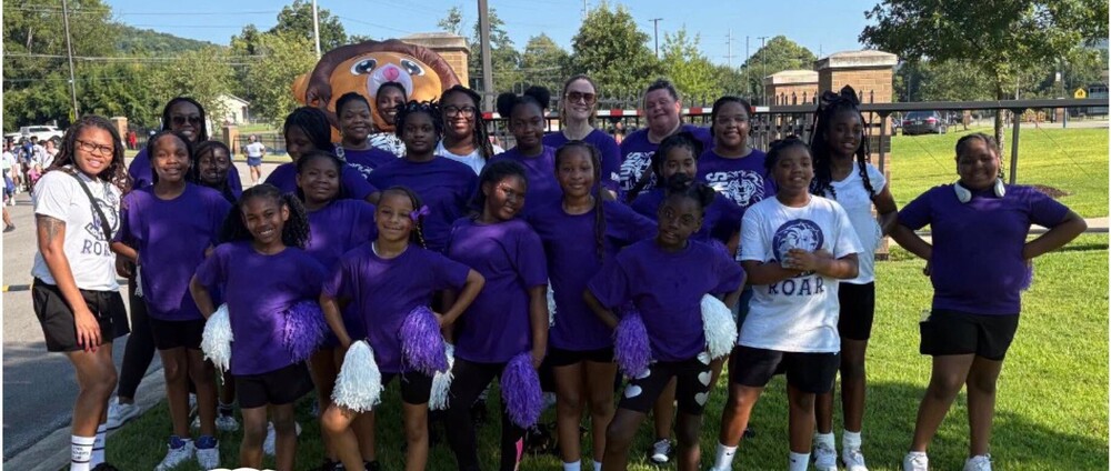 A large group of girls are dressed in purple shirts and holding white and purple pompoms.  They are standing outside.