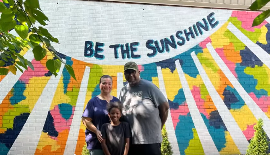 2 Grandparents and their granddaughter are standing outside in front of a large mural that says "Be the Sunshine"