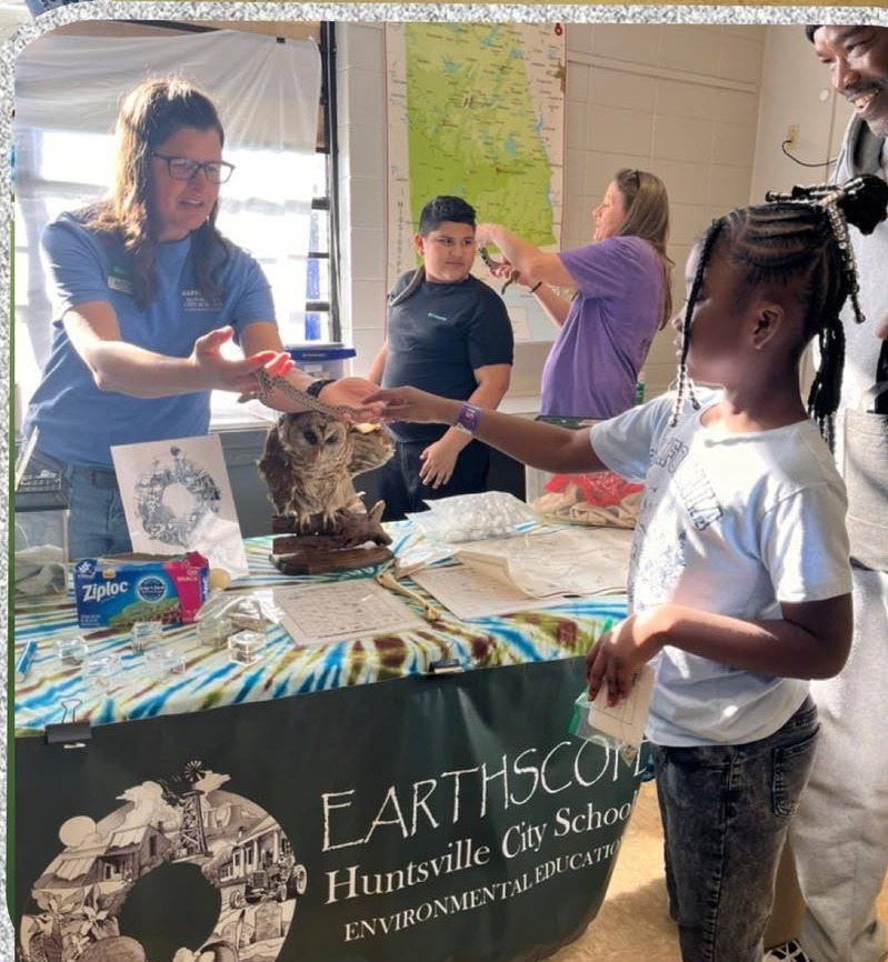 A young girl is standing at a table with a covering that says "Earthscope" and she's touching an owl on the table.  A woman is standing at the table with her.