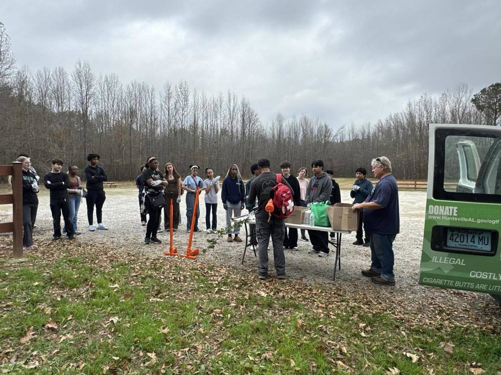 A group of Lee High School students stands outdoors at Hayes Nature Preserve with members of the City of Huntsville’s Green Team. The students are gathered in a semicircle around a table where tools and supplies are laid out, listening to instructions before beginning invasive species removal. Two orange weed pullers stand upright in the foreground, and a Green Team vehicle is parked to the right. Bare trees and cloudy skies appear in the background.