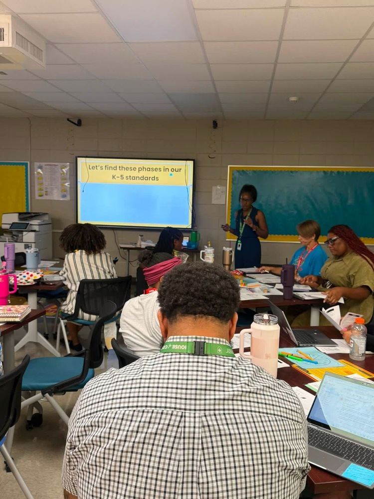 Several teachers in a room viewing a monitor with yellow and blue background.