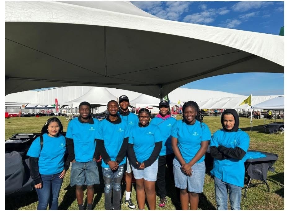 8 students from JROTC wearing blue tshirts with black long sleeved shirts under them standing under white tent at the Toyota fiield