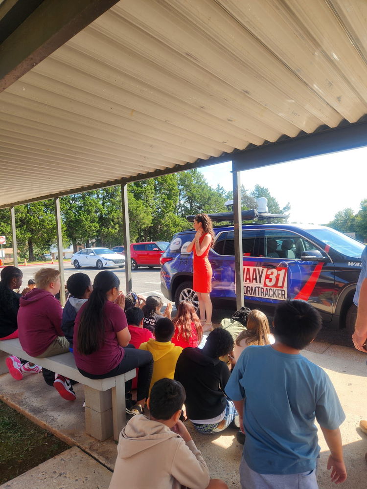 A group of students sits outside listening to WAAY 31 meteorologist Grace Anello speak next to the WAAY 31 StormTracker vehicle.