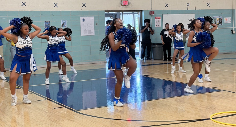 Cheerleaders dressed in blue and white cheer uniforms perform a synchronized routine holding blue pom-poms on the gymnasium court floor.