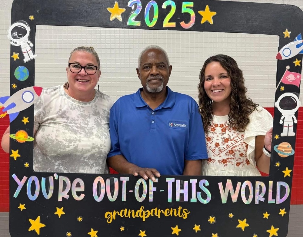 Mrs. Ogle and Ms. Bodkin with their honorary grandparent, Mr. Leon, pose behind a space-themed frame with the text You're Out of This World Grandparents 2025.