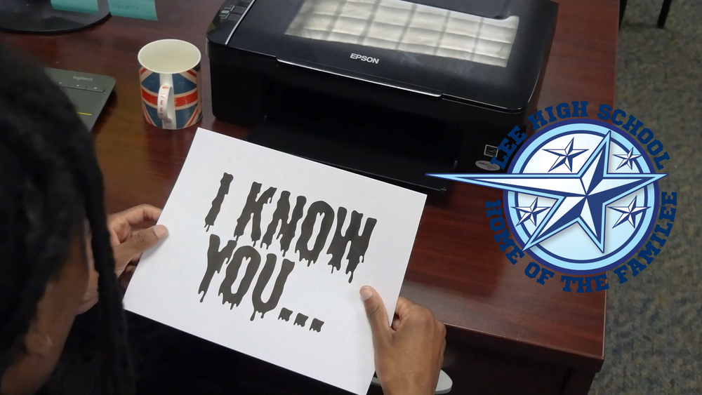 Student holds a printed sheet reading “I KNOW YOU…” in dripping black letters beside an Epson printer on a desk, with the Lee High School “Home of the Familee” logo displayed on the right side of the image.