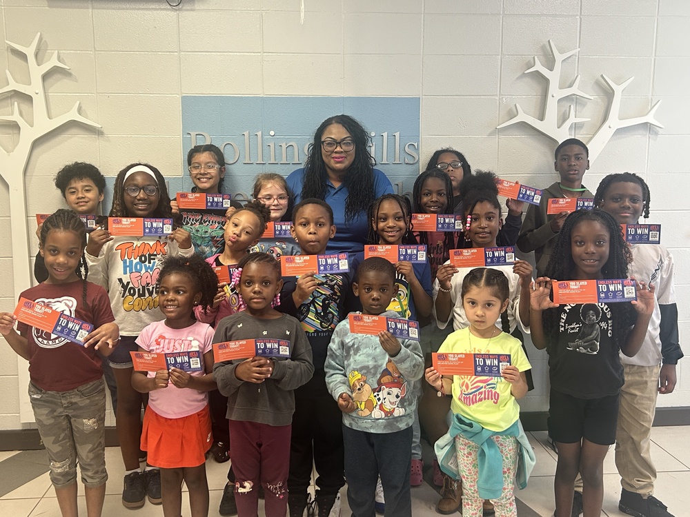 A group of Rolling Hills Elementary students pose proudly with their “Student of the Month” certificates from Dave & Buster’s. They are smiling and holding up their prizes while standing in front of a wall with the school’s name and logo. Principal Mrs. Agun stands in the center behind the students, smiling with them.