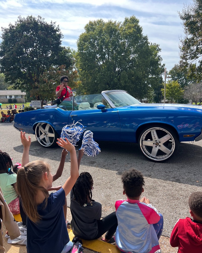 Dr. Headen rides in a blue car leading the Hispanic Heritage Parade while students sit nearby and celebrate the event.