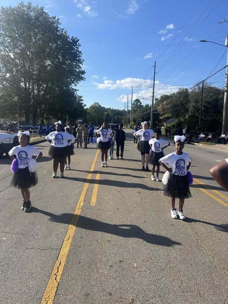 A group of cheerleaders are dressed in white Dawson shirts with black skirts. They have purple and white pom-poms and are lined up in the street for the parade.