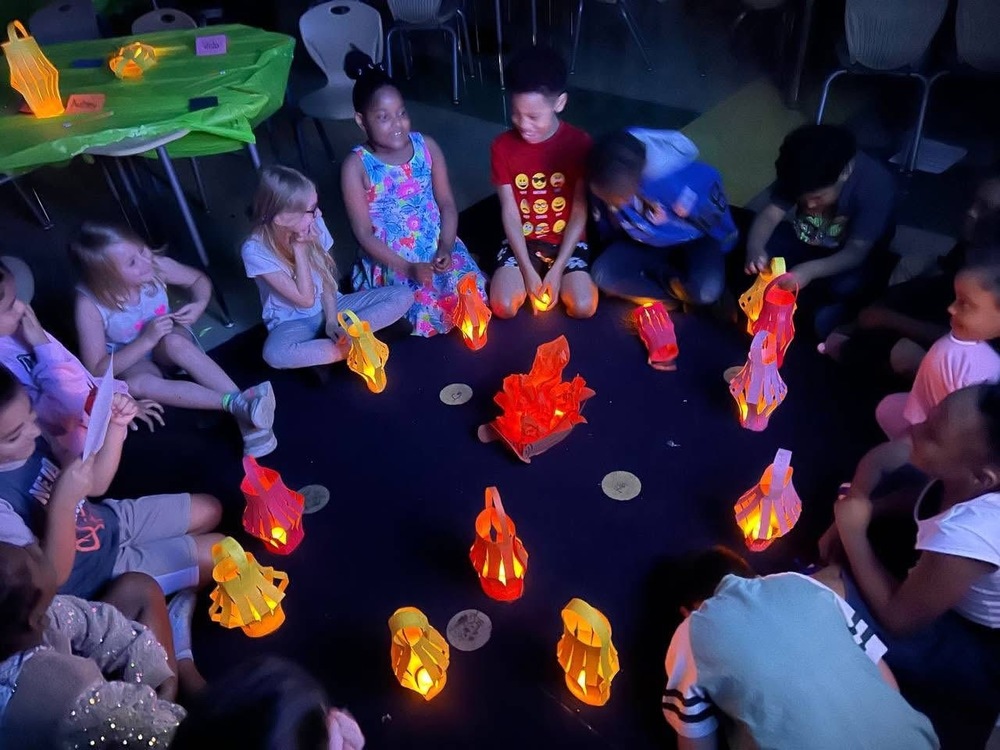 Students in a circle, seating in the dark, with red and yellow objects for camping room transformation