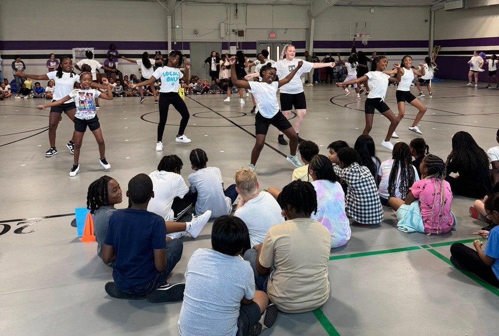 A group of 7 girls in white shirts with black shorts are dancing in front of a large group of students in the gymnasium