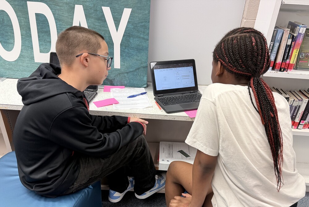 Two students sit side-by-side on blue ottomans at a counter, collaborating while looking at an open laptop screen in a classroom next to a bookshelf.