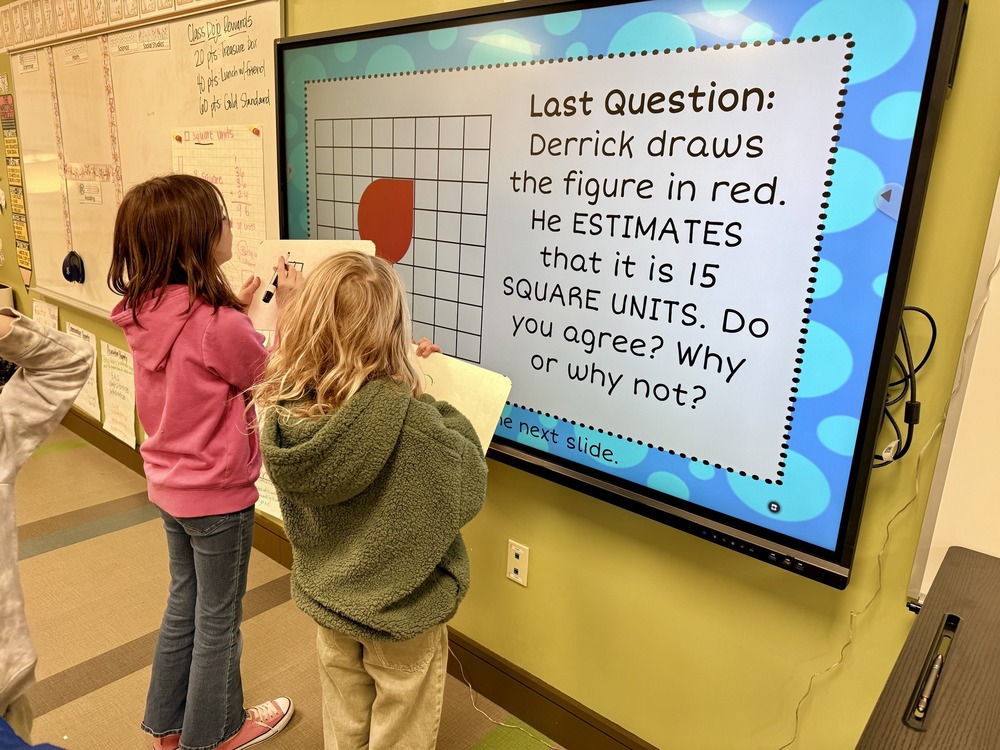 Two students are in front of the board working on the last question "Derrick draws the figure in red. He estimates that it is 15 square units. Do you agree? Why or why not?"