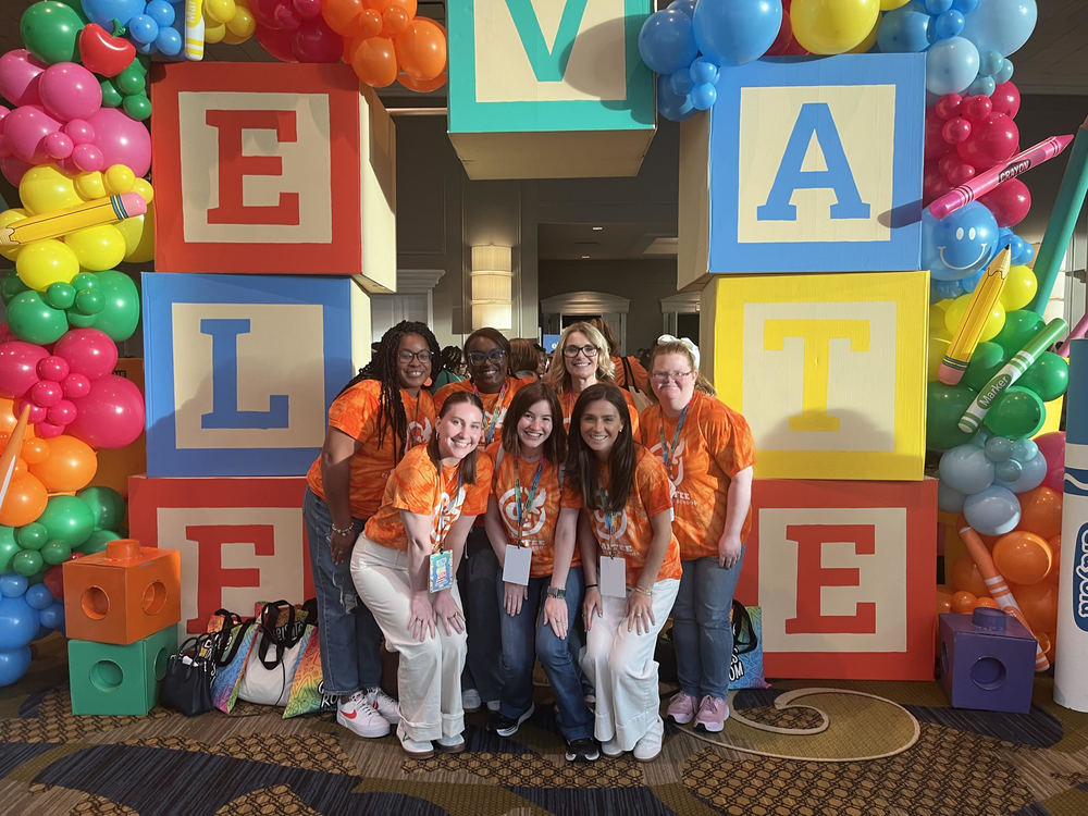 Ms. Fudge, Ms. Spencer, Ms. Honda, Ms, Wall, Ms, Bartley, Ms. Haynes, and Ms. O'Connor standing in front of an ELEVATE sign. They are at the ELEVATE conference wearing orange chaffee shirts
