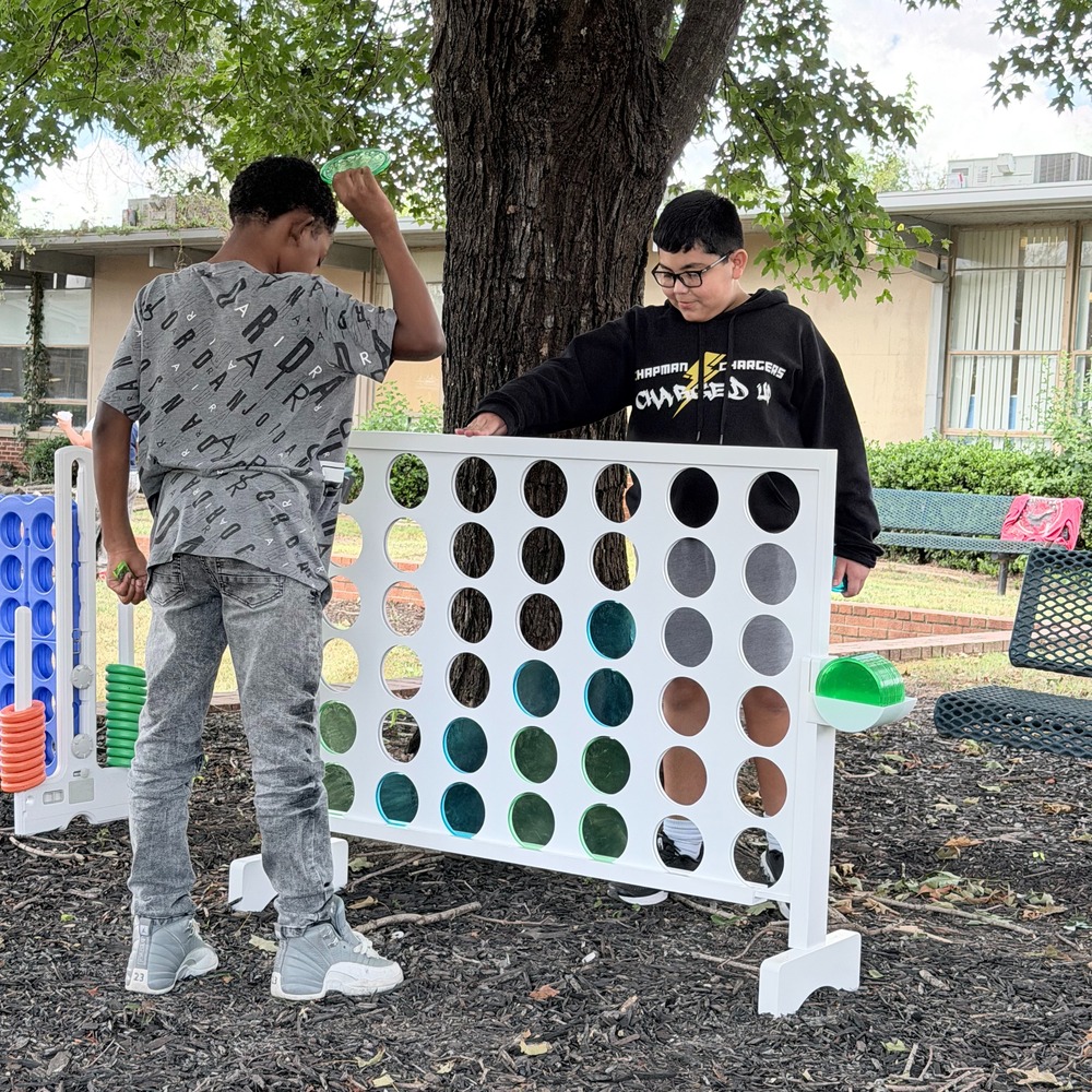 Two middle school students play an oversized white wooden Connect Four game outdoors under a large shade tree.