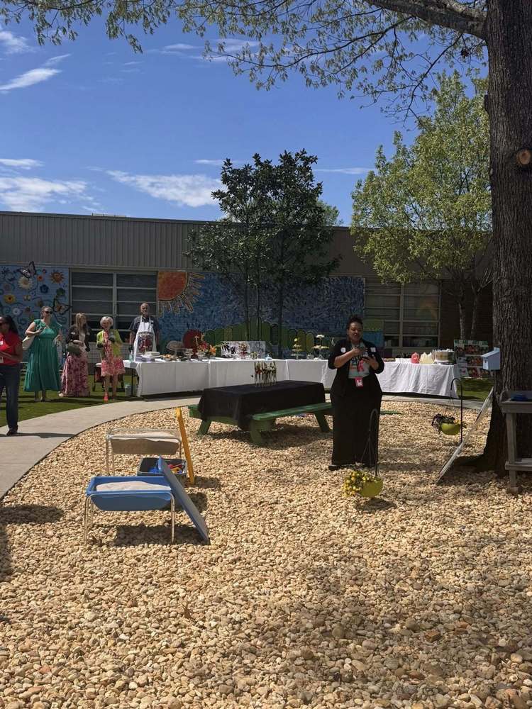 A group is standing outside in a courtyard listening to a speaker .  They are surrounded by trees, a table of food, and sand play tables.