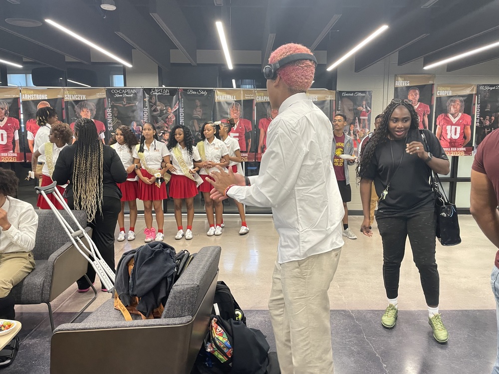 The cheer team in red and white uniforms with cheer coach in front of them wearing black , football team member with pink hair and headphones looking at the cheerleaders with athletic director in all black and gray chairs with a pair of crutches can be seen