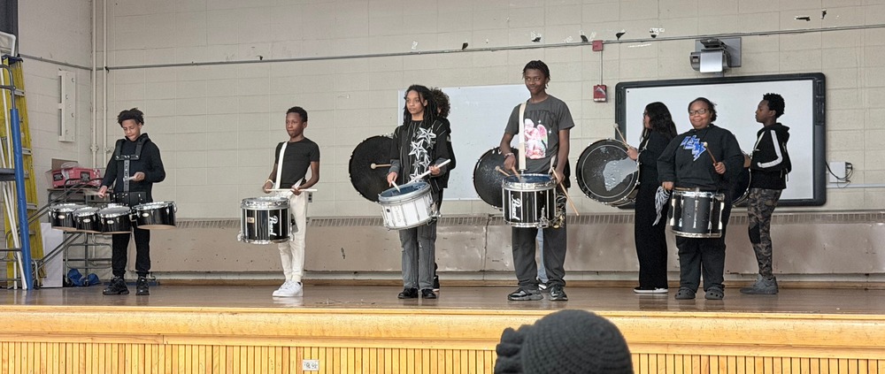 A group of Chapman Middle School students from the drumline performs on a wooden stage in a gym. They are playing various drums during the school’s talent show.