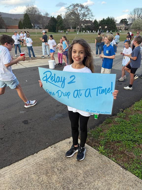 5th grade student holding a blue Run for Water relay sign with student runners in the background.