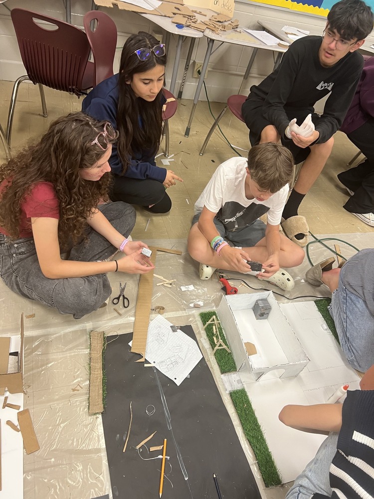 Four students sitting on the ground, gluing pieces together to build pieces for their house which is part of their math project
