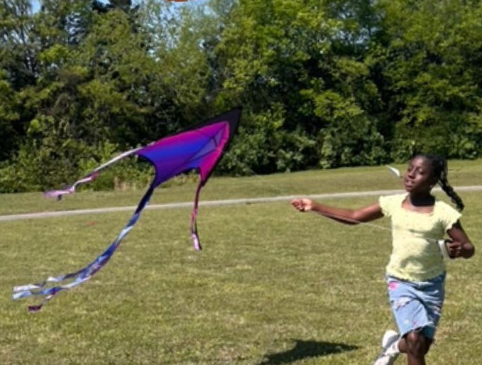 A girl is running through a field holding a pink and blue kite behind her.