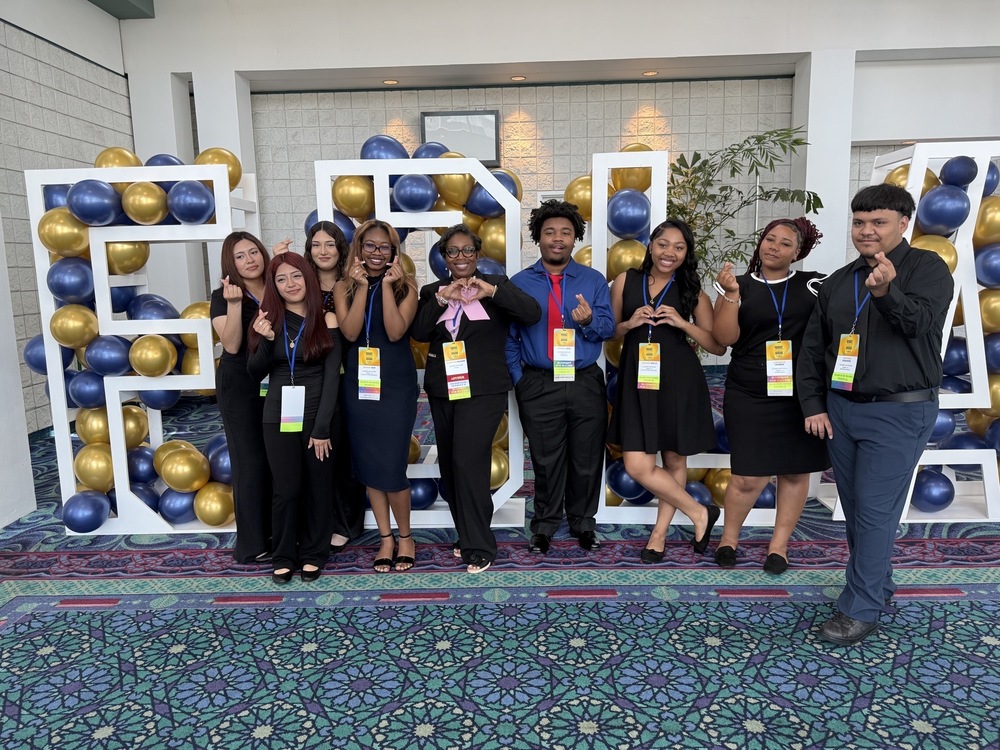 8 students of the Future Business Leaders of America dressed in dark blue and black posing for picture with Adviser standing in the middle with Large White Lettere, FBLA with blue and gold balloons