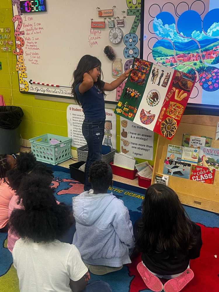 A young girl stands in front of a classroom showing a colorful poster with images representing Hispanic culture, including food, people, and celebrations. Several classmates sit on a rug, attentively watching her presentation. The classroom walls are decorated with learning charts, a whiteboard, and bright educational materials.