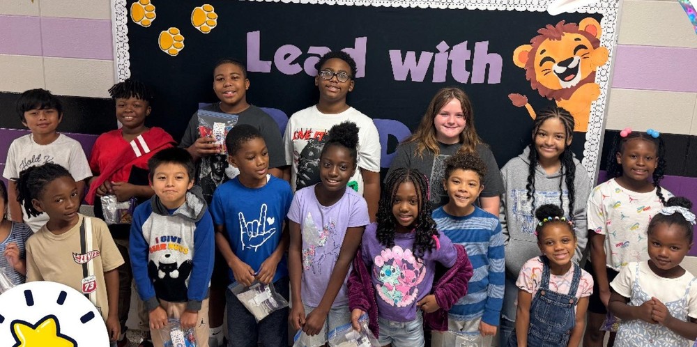 A group of 15 students are standing in front of a black bulletin board that says "Lead with PRIDE" and has a lion clipart on the top right, with paw prints on the top left.