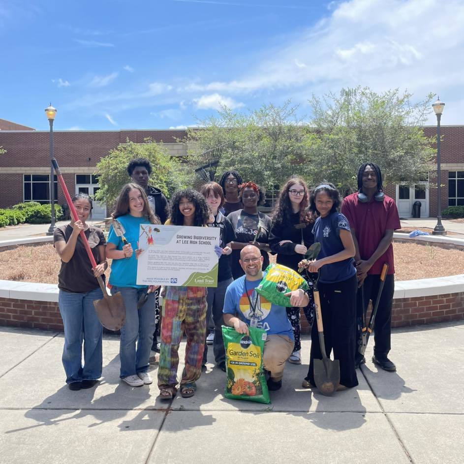 Members of the Green Ambassadors team stand together in Lee High School’s theater courtyard holding a large sign celebrating their biodiversity initiative. Several students hold shovels used during the project. Mr. McDonald kneels in front of the group, smiling and holding two bags of garden soil. The circular courtyard planter and school building are visible behind them on a bright, clear day.