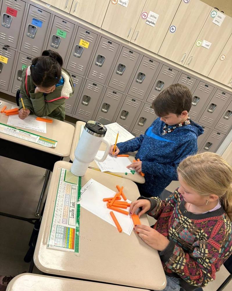 Three students use base ten blocks on their desks to solve a math problem.