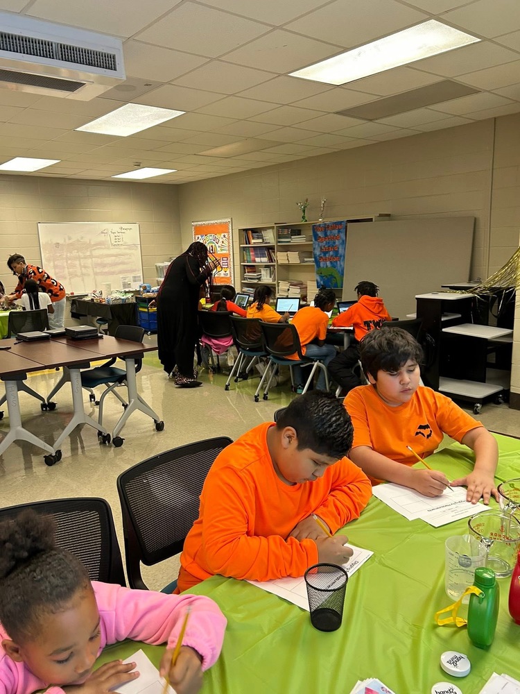 2 male students pictured with 1 girl working on a green table cloth. Other students are working in small group along with 4th grade teachers
