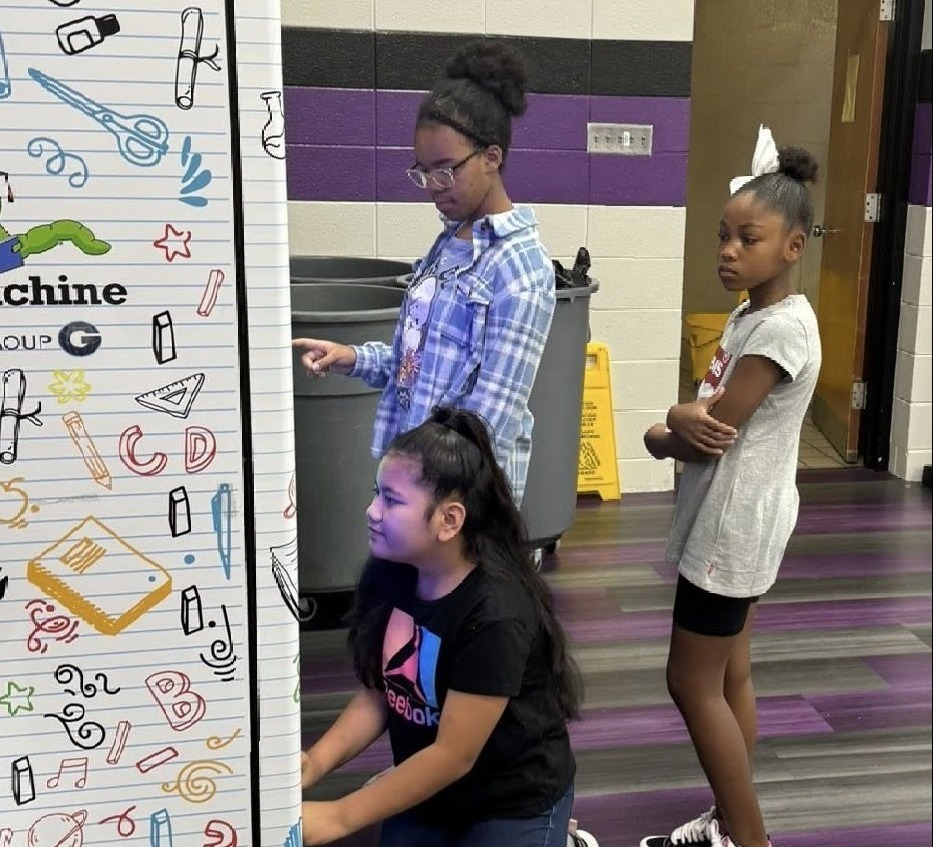 Three girls are in a school cafeteria at a book vending machine. One girl is crouched down getting a book out of the slot, while the other two girls are standing behind her waiting.