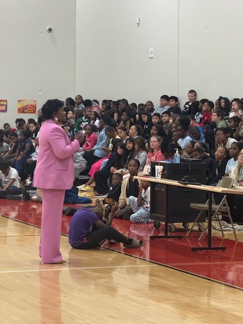 Principal Walters, dressed in a pink suit, stands on the gym floor speaking into a microphone during an ACAP pep rally. A large group of students sit closely together on bleachers and along the floor, attentively listening. The setting is a school gymnasium, with a table holding audio equipment nearby, as the student body gathers to be motivated and energized for testing.