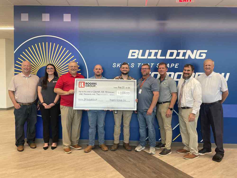 Picture of teachers from the Huntsville Center of Technology holding a check for $1,000 along side members from The Rogers Group. They are standing in front of a wall in the Huntsville Center for Technology that says Building skills that shape tomorrow.