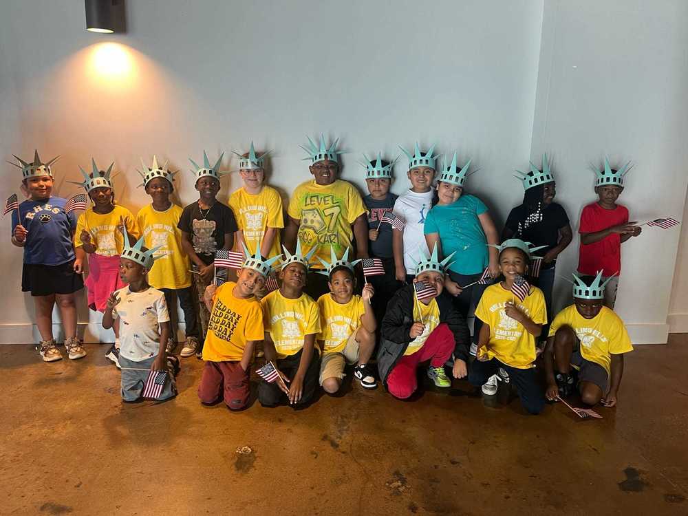 A group of 2nd grade students wearing Statue of Liberty paper crowns and holding American flags pose together, many in yellow “Rolling Hills Elementary Field Day” shirts.