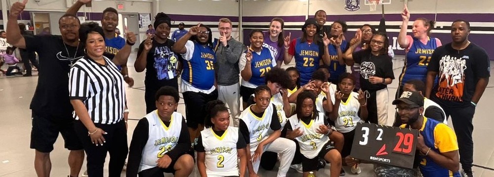 A group of adults and students are standing in the gym.  The adults are wearing blue and yellow Jemison  jerseys .  The students are seated on the ground in white and yellow Jemison jerseys.  Another teacher is wearing a referee shirt standing with the two teams.