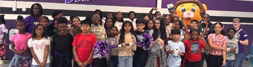 A large group of students are standing together in the school gym.  They are holding a large stick with purple pompoms at each end. 