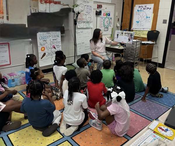 A teacher is sitting in a classroom with a group of students gathered around her on the carpet. She is reading a book to the students.