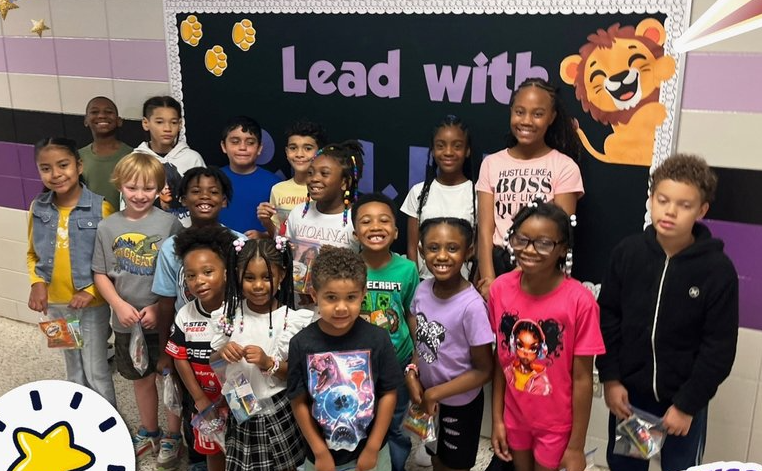 A group of 18 students, ranging from Kindergarten to 5th grade are posed in front of a black bulletin board that says "Lead with PRIDE" in purple letters with a lion cub clipart in the corner.  